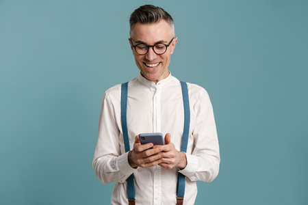 Happy grey-haired man in eyeglasses smiling and using cellphone isolated over blue backgroundの写真素材