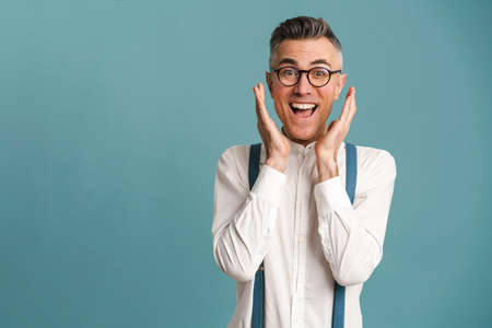 Excited grey-haired man in eyeglasses expressing surprise on camera isolated over blue background