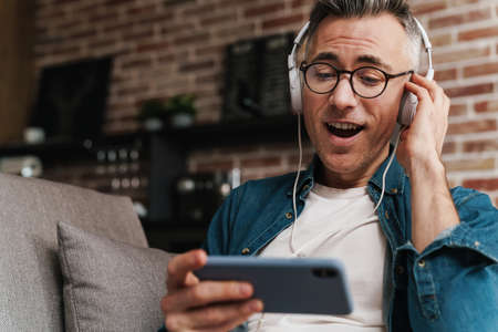 Excited man in headphones using mobile phone while sitting on sofa at homeの写真素材