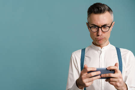 Shocked grey-haired man in eyeglasses playing online game on cellphone isolated over blue background