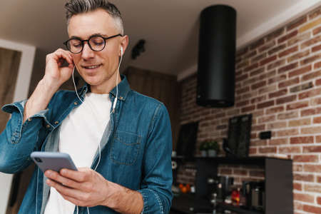 Cheerful white-haired man in eyeglasses using earphones and mobile phone at homeの写真素材