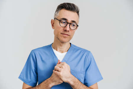 Sad white-haired medical doctor posing and holding hands together isolated over white wall