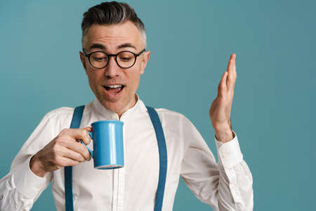 Surprised grey-haired man in eyeglasses exclaiming while drinking coffee isolated over blue backgroundの写真素材