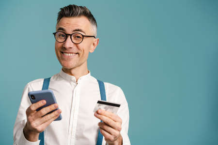 Happy grey-haired man in eyeglasses posing with cellphone and credit card isolated over blue...