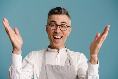 Delighted grey-haired man wearing apron exclaiming with hands up isolated over blue background