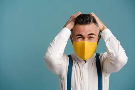 Displeased grey-haired man in face mask posing with grabbing his head isolated over blue background