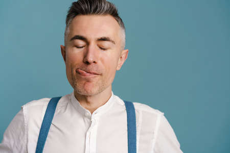 Happy grey-haired man liking his lips while posing at camera isolated over blue background