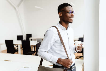 Afro american pleased man in eyeglasses smiling and drinking coffee in officeの写真素材