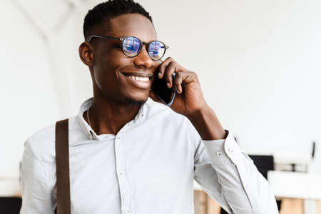 Afro american pleased man in eyeglasses smiling and talking on mobile phone in office