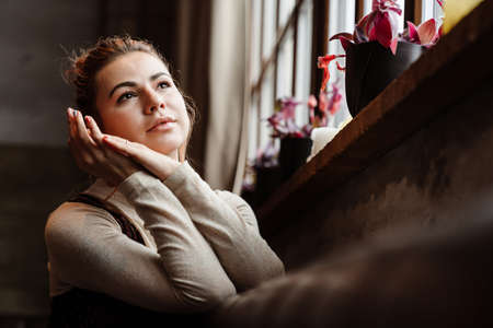 Young caucasian beautiful woman looking aside while sitting by window indoors