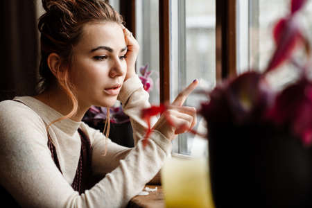 Young caucasian beautiful woman drawing on window glass while sitting indoors