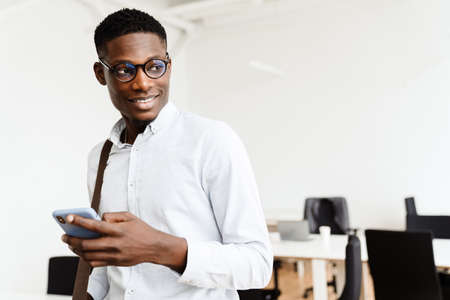 Afro american pleased man in eyeglasses smiling and using mobile phone in officeの写真素材