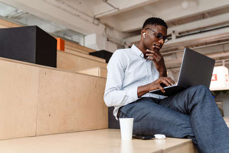 Afro american focused man in earphones working with laptop while sitting indoors