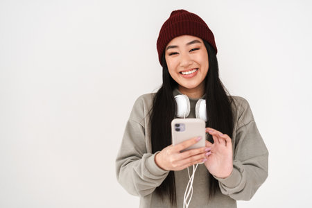 Asian joyful nice girl with headphones using mobile phone isolated over white backgroundの写真素材
