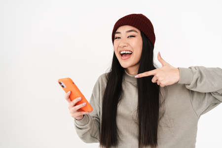 Asian smiling girl holding and pointing finger at mobile phone isolated over white background