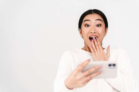 Asian surprised nice girl playing online game on mobile phone isolated over white backgroundの写真素材