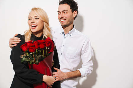 Lovely romantic young happy couple celebrating an event together, woman holding bouquet of red roses isolated over white backgroundの写真素材