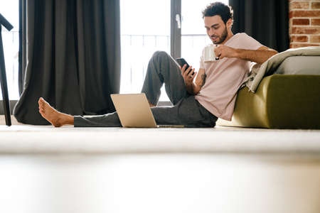 Handsome pleased man using mobile phone and drinking coffee while sitting on floor in bedroom