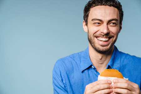 Cheerful handsome guy laughing while posing with hamburger isolated over blue background