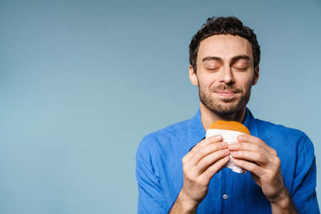 Cheerful handsome guy smiling while posing with hamburger isolated over blue backgroundの写真素材