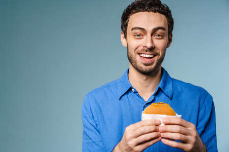 Joyful handsome guy smiling while posing with hamburger isolated over blue background