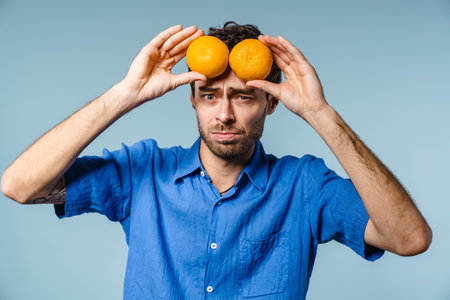 Confused handsome unshaven guy posing with mandarins isolated over blue backgroundの写真素材