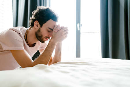 Focused unshaven man praying while holding hands together in bedroom at home