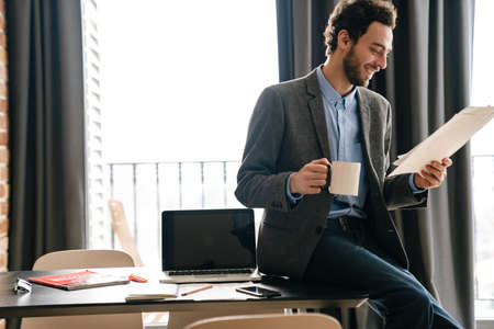 Handsome joyful businessman smiling and examining documents while drinking coffee indoorsの写真素材