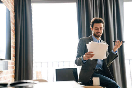 Handsome pleased businessman talking on mobile phone while examining documents indoors