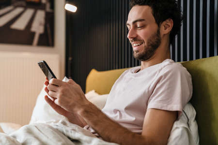 Joyful brunette man smiling and using mobile phone while lying in bedの写真素材