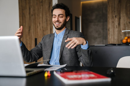 Handsome joyful businessman gesturing while working with laptop at homeの写真素材