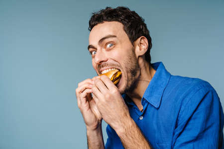 Excited handsome hungry guy eating hamburger on camera isolated over blue backgroundの写真素材