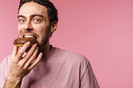 Joyful handsome hungry guy eating doughnut on camera isolated over pink background