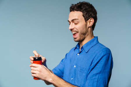 Joyful handsome guy smiling while opening soda can isolated over blue backgroundの写真素材
