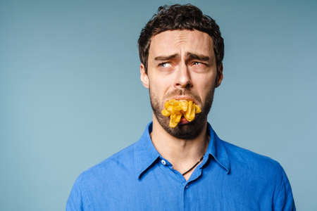 Unhappy handsome guy posing with french fries in his mouth isolated over blue background