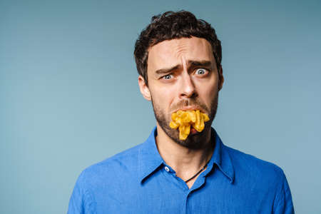 Unhappy handsome guy posing with french fries in his mouth isolated over blue backgroundの写真素材