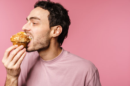 Happy handsome guy eating muffin with eyes closed isolated over pink background
