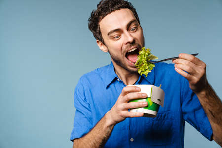 Excited handsome hungry guy eating salad takeaway isolated over blue backgroundの写真素材
