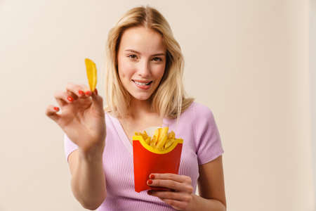 Cheerful beautiful girl smiling while showing french fries at camera isolated over white backgroundの写真素材