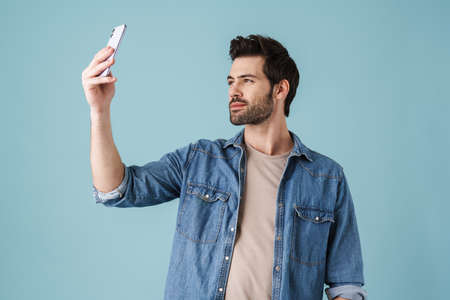 Young handsome unshaven man smiling and looking at cellphone isolated over blue background