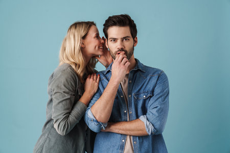 Young beautiful woman whispering secret at surprised guy isolated over blue backgroundの写真素材