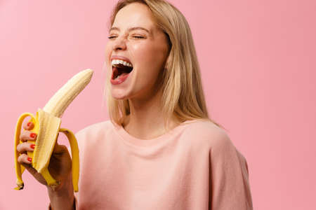 Joyful charming blonde girl laughing and eating banana isolated over pink backgroundの写真素材