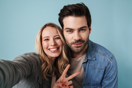 Young happy couple gesturing peace sign while taking selfie photo isolated over blue backgroundの写真素材