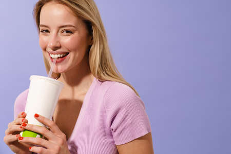 Happy charming blonde girl smiling while drinking soda isolated over blue background