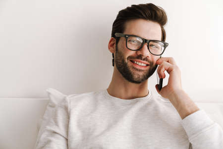Smiling young man talking on mobile phone while resting in bed at homeの写真素材