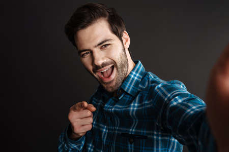 Joyful handsome guy pointing finger at camera while taking selfie isolated over black background