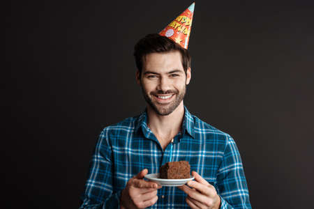 Smiling guy in party cone posing with birthday cake isolated over black backgroundの写真素材