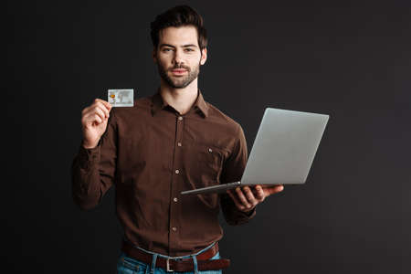 Confident unshaven guy posing with credit card and laptop isolated over black backgroundの写真素材