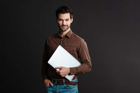 Pleased handsome brunette guy smiling while posing with laptop isolated over black background