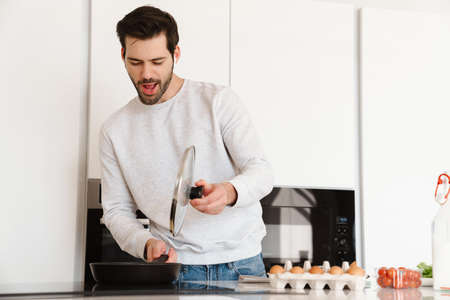 Happy young man using earphones while cooking scrambled eggs at home kitchen